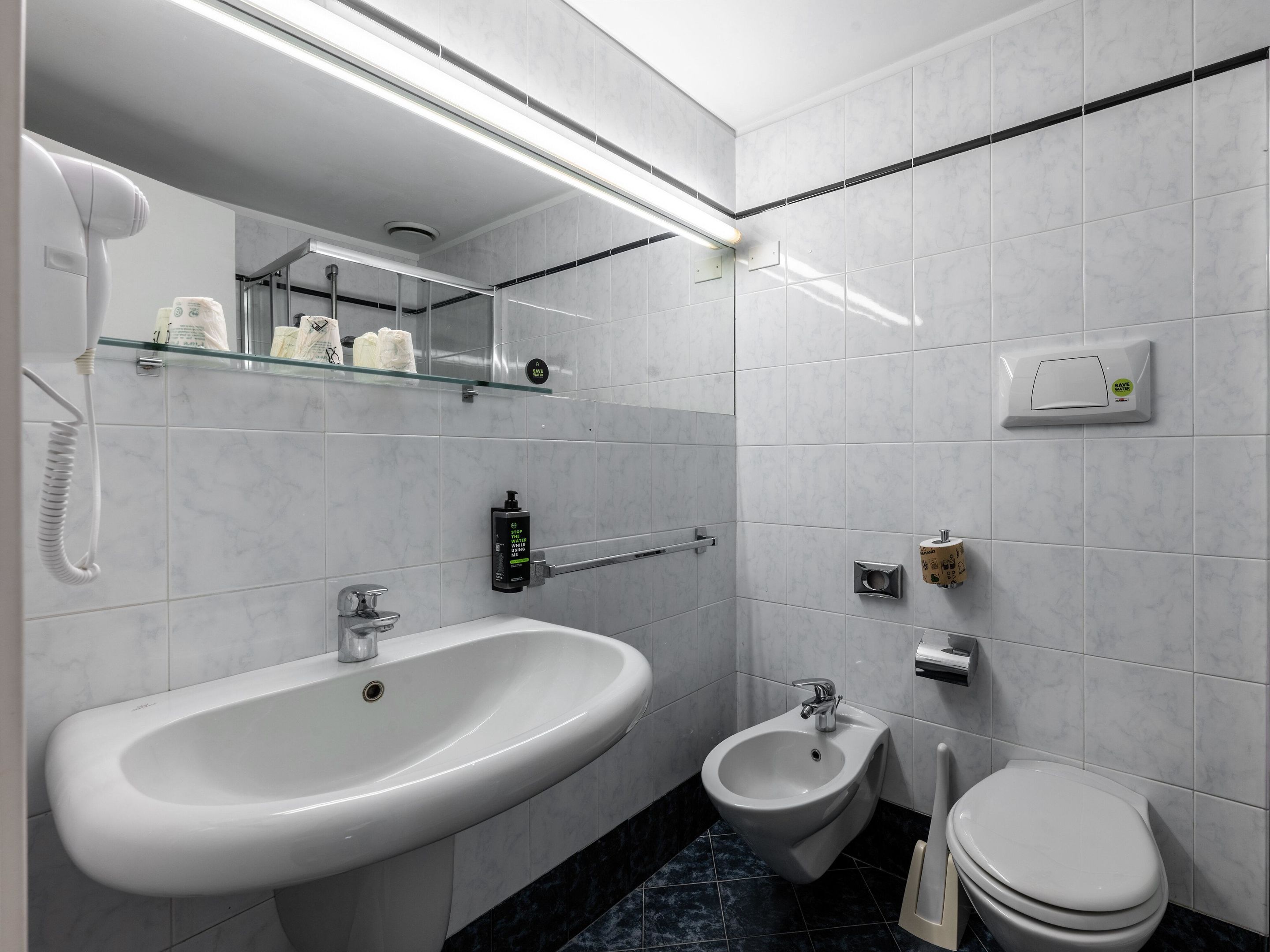 A modern bathroom with a sink, bidet, toilet, and a glass shelf with toiletries.