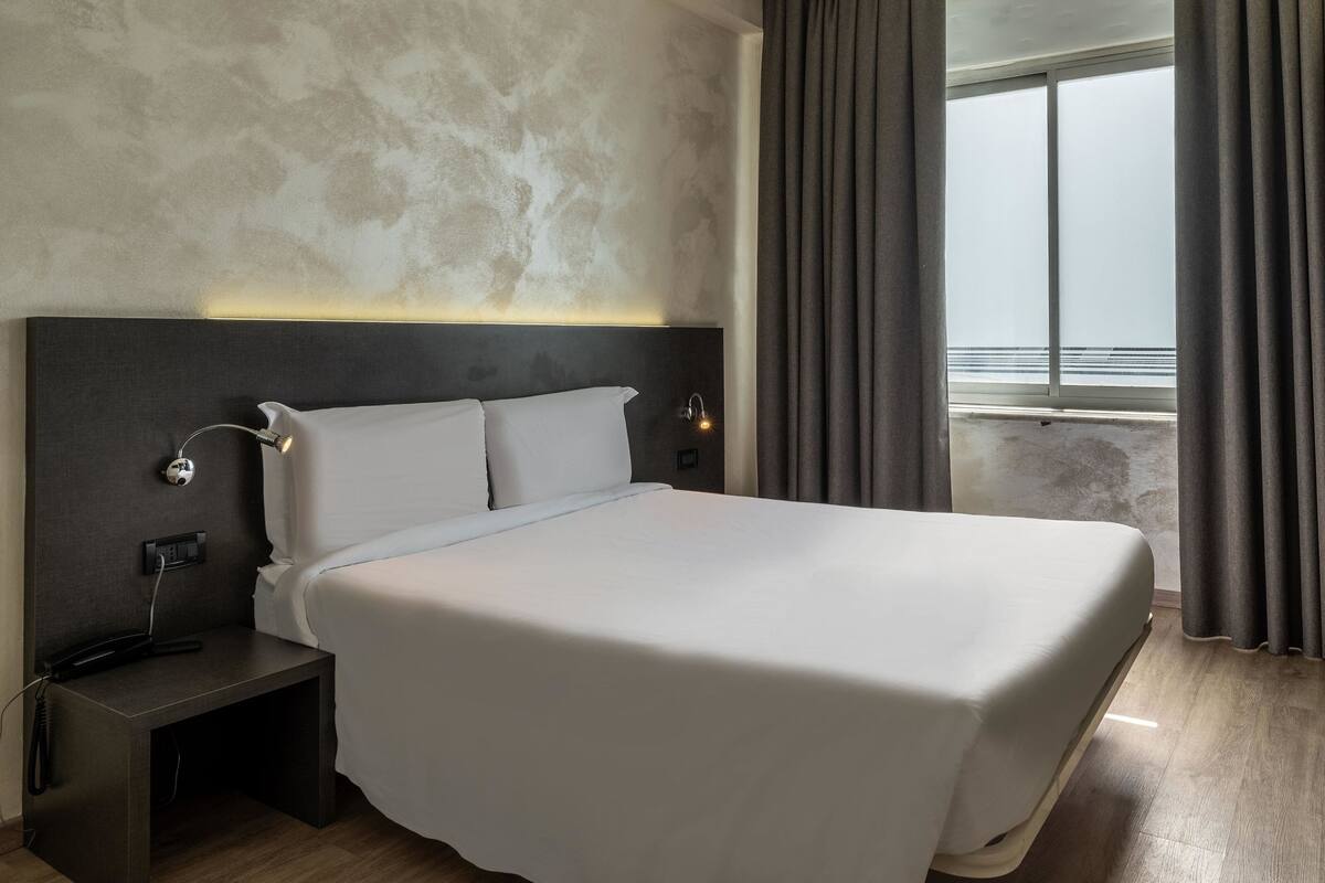 A modern hotel room with a large bed, a bedside table with a lamp, and a view of the sea through a window.