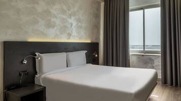 Una habitación de hotel moderna con una cama grande, una mesita de noche con lámpara y vistas al mar a través de una ventana.