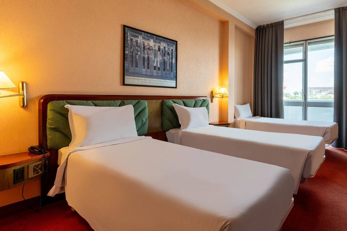 A hotel room with two beds, a painting on the wall, and a window with curtains.