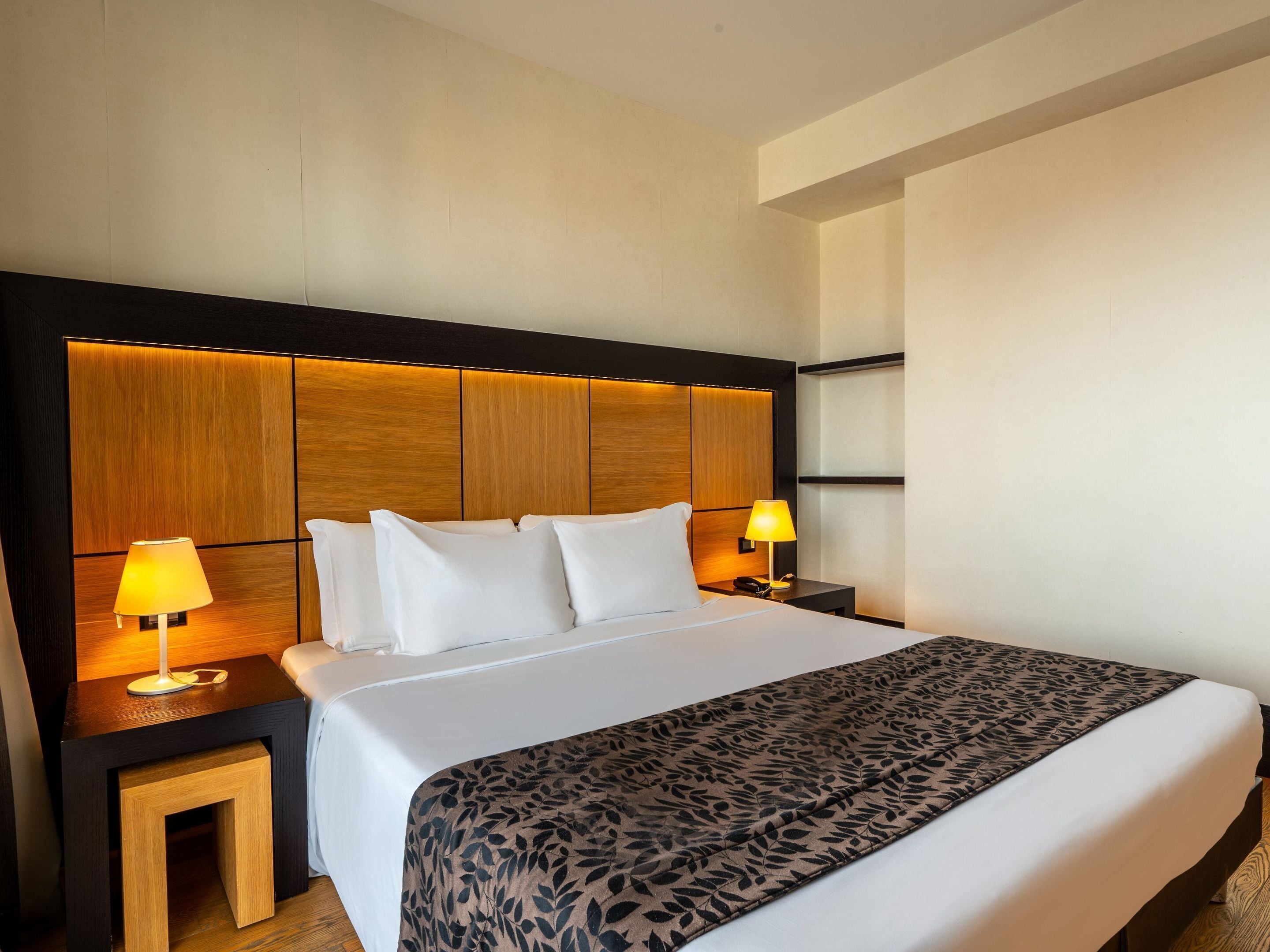 A hotel room with a large bed, two bedside tables with lamps, a wooden headboard, and a patterned throw blanket.