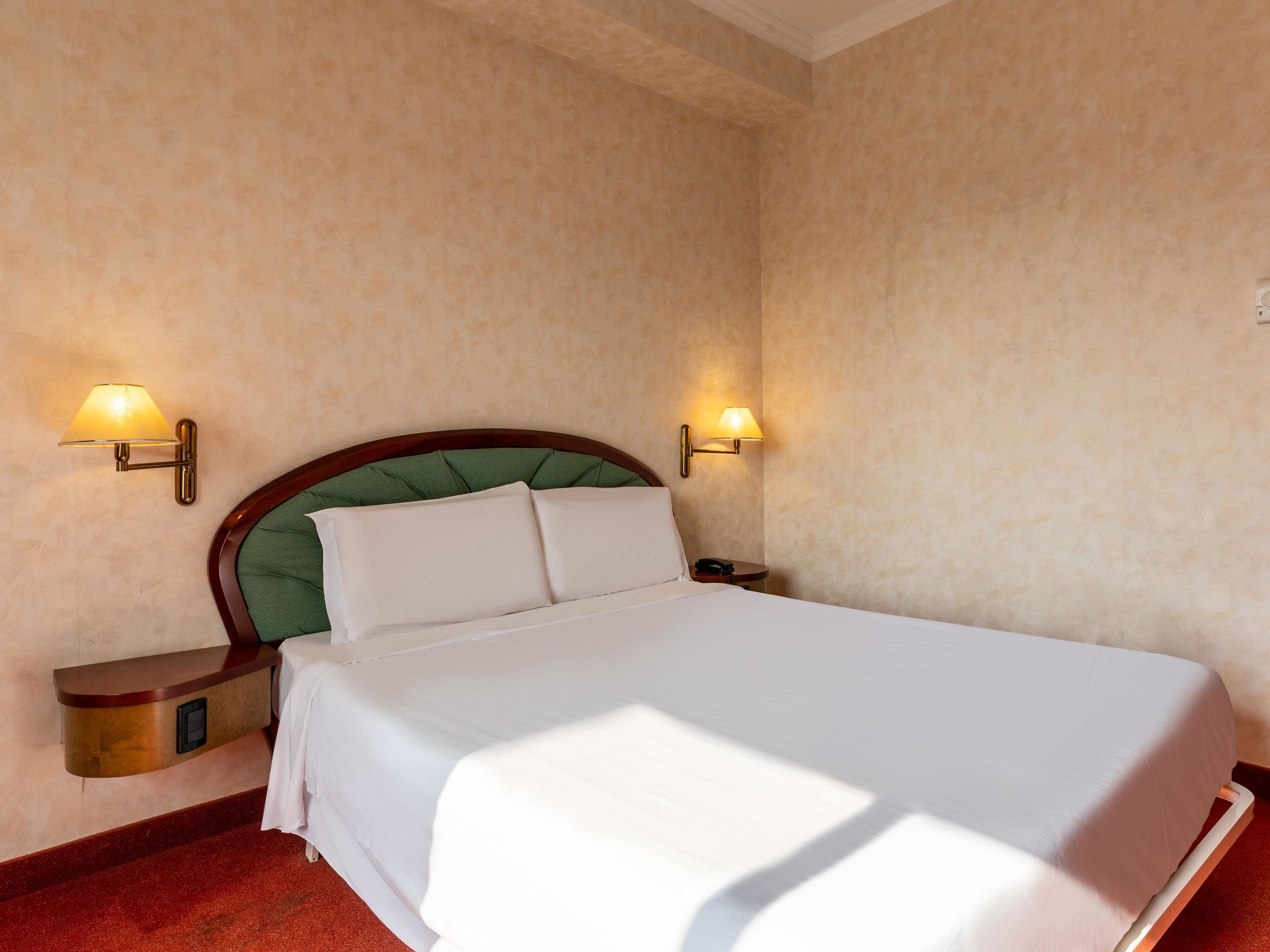 A neatly made bed with white linens, a green upholstered headboard, and two bedside lamps on either side.