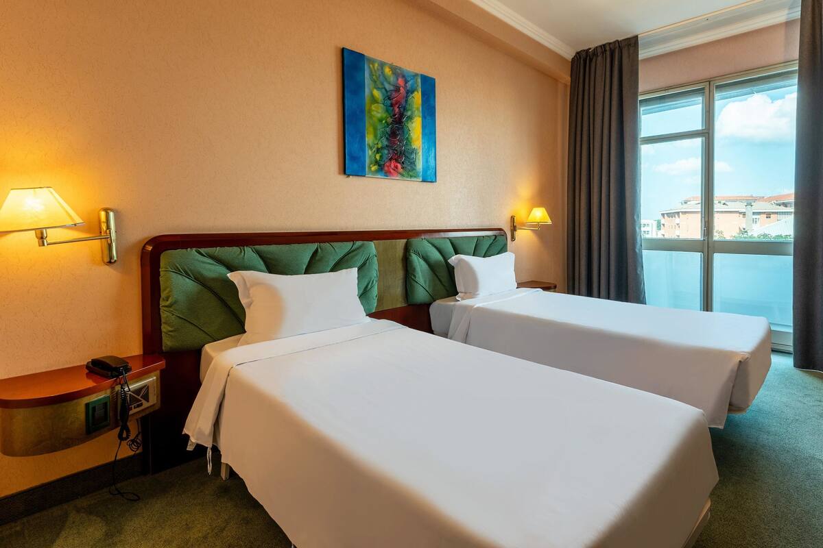 A hotel room with two beds, a painting on the wall, and a window with a view of buildings.