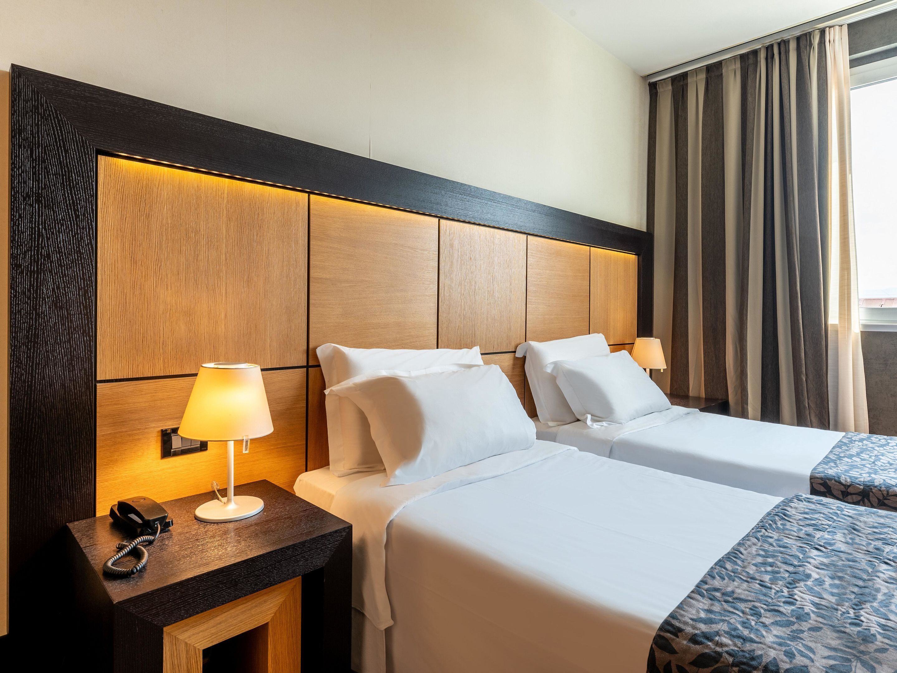 A hotel room with two beds, a wooden headboard, bedside lamps, a telephone, and a window with curtains.