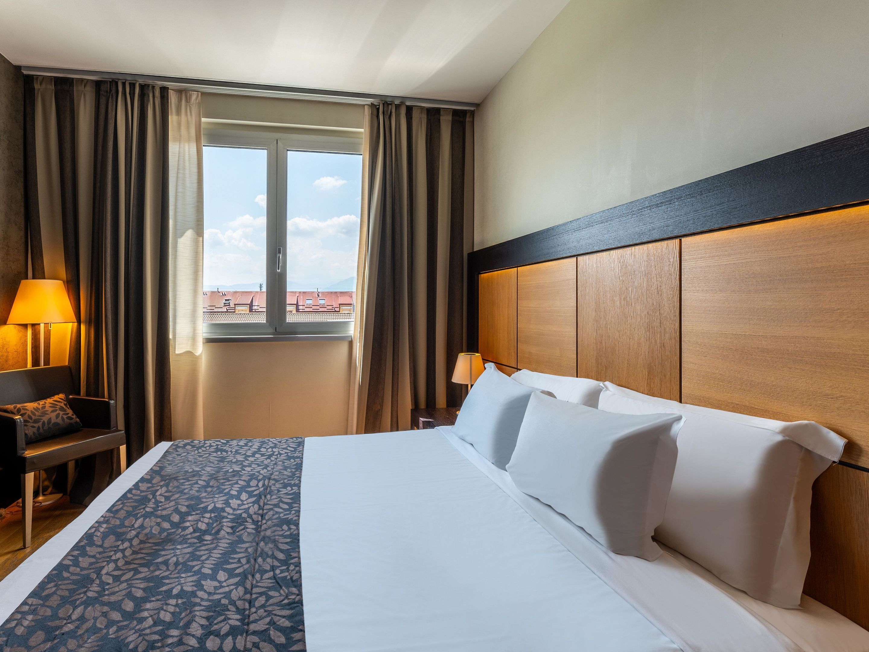 Een hotelkamer met een bed, een bureau, een stoel, een lamp en een raam met gordijnen.