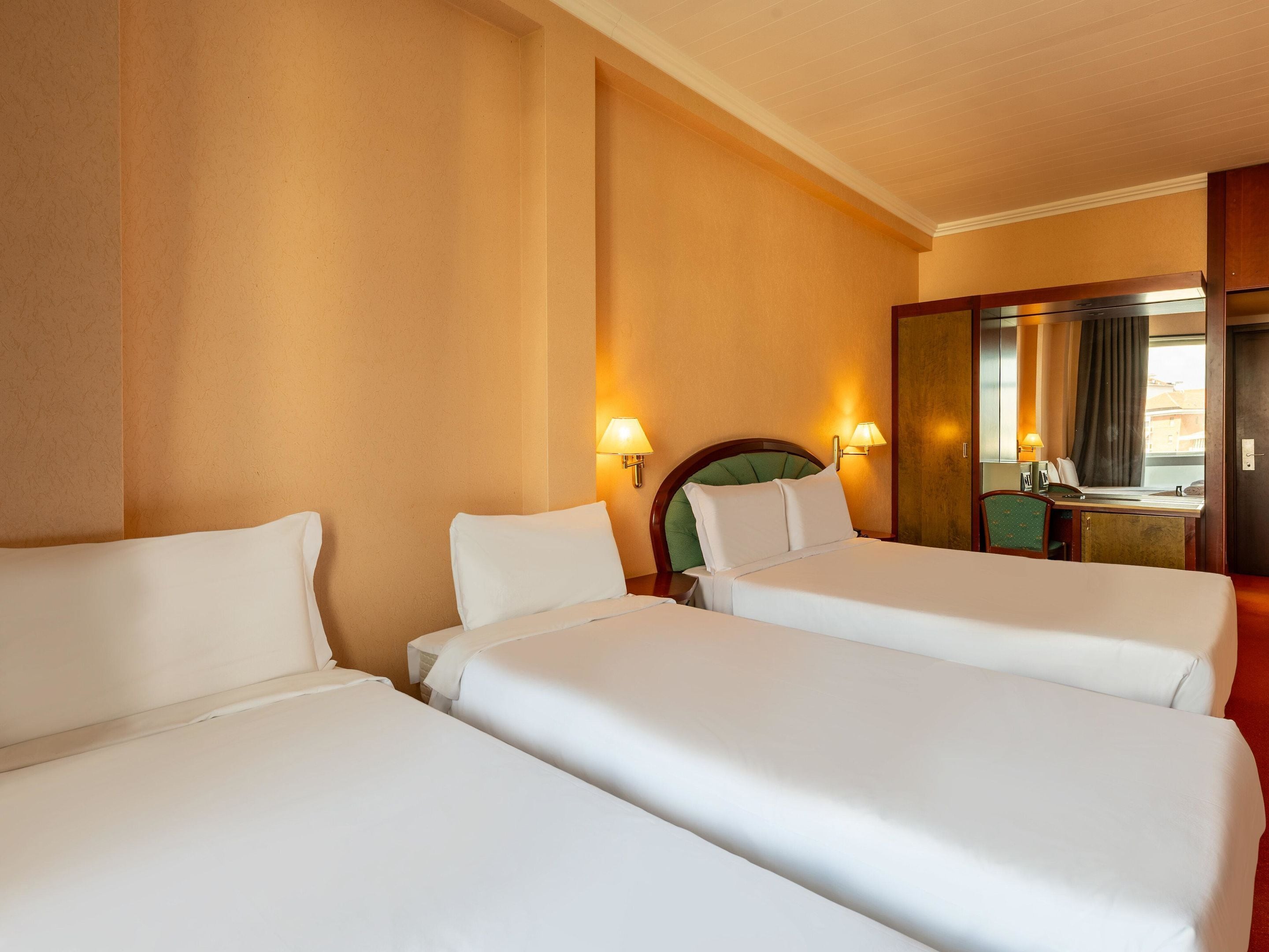 A hotel room with two single beds, a desk, a chair, a mirror, and two wall-mounted lamps.