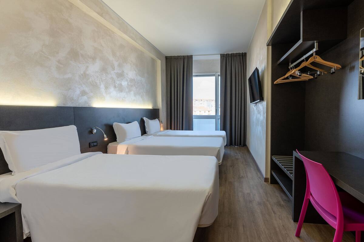 A hotel room with two beds, a desk, a TV, and a view of the city.