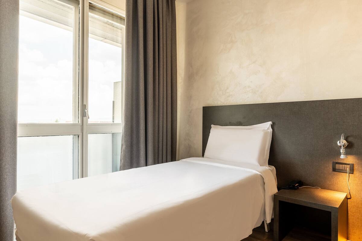 A hotel room with a large bed, a nightstand with a lamp, and a window with curtains.