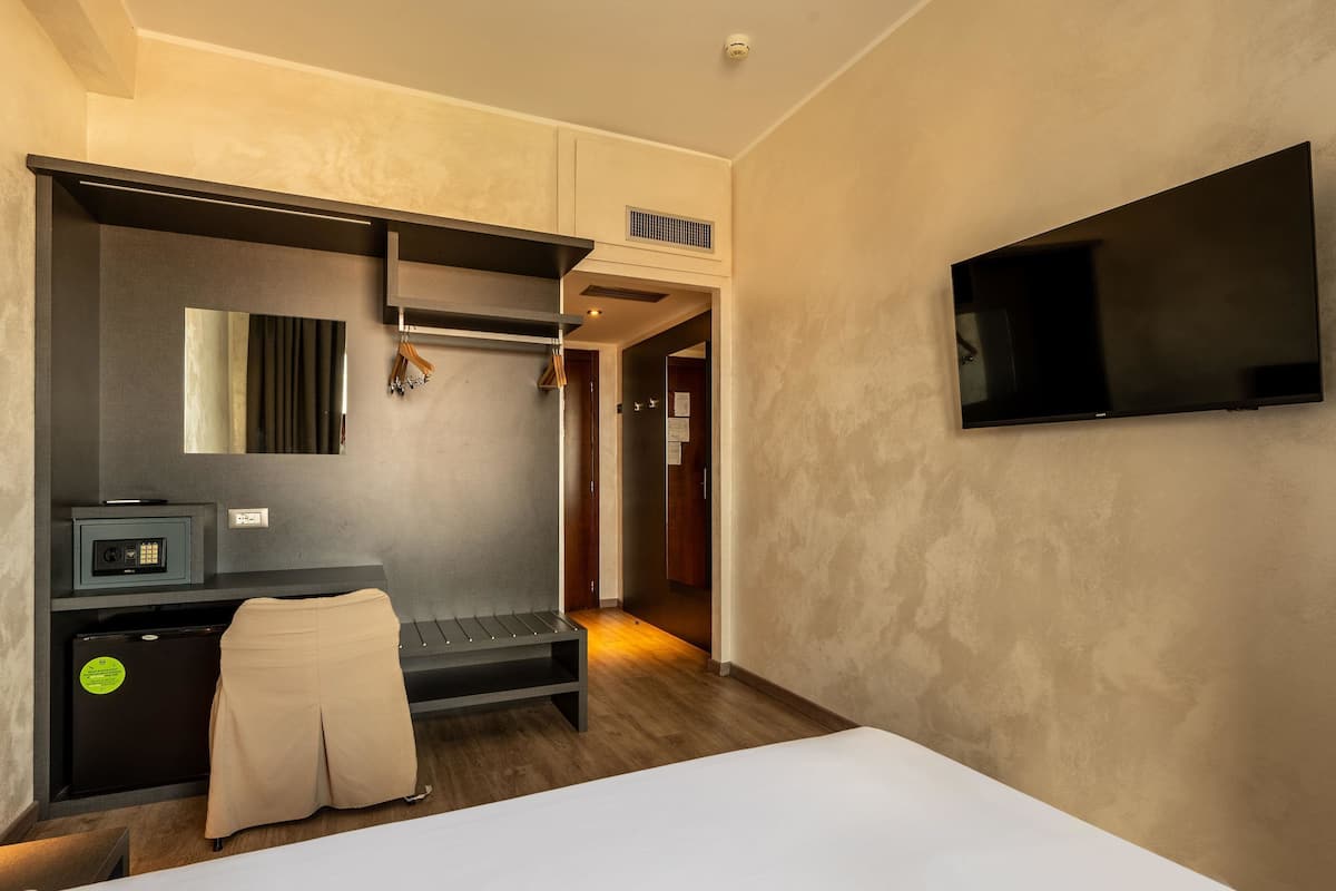A hotel room with a bed, a desk with a computer, a TV mounted on the wall, and a coat rack.