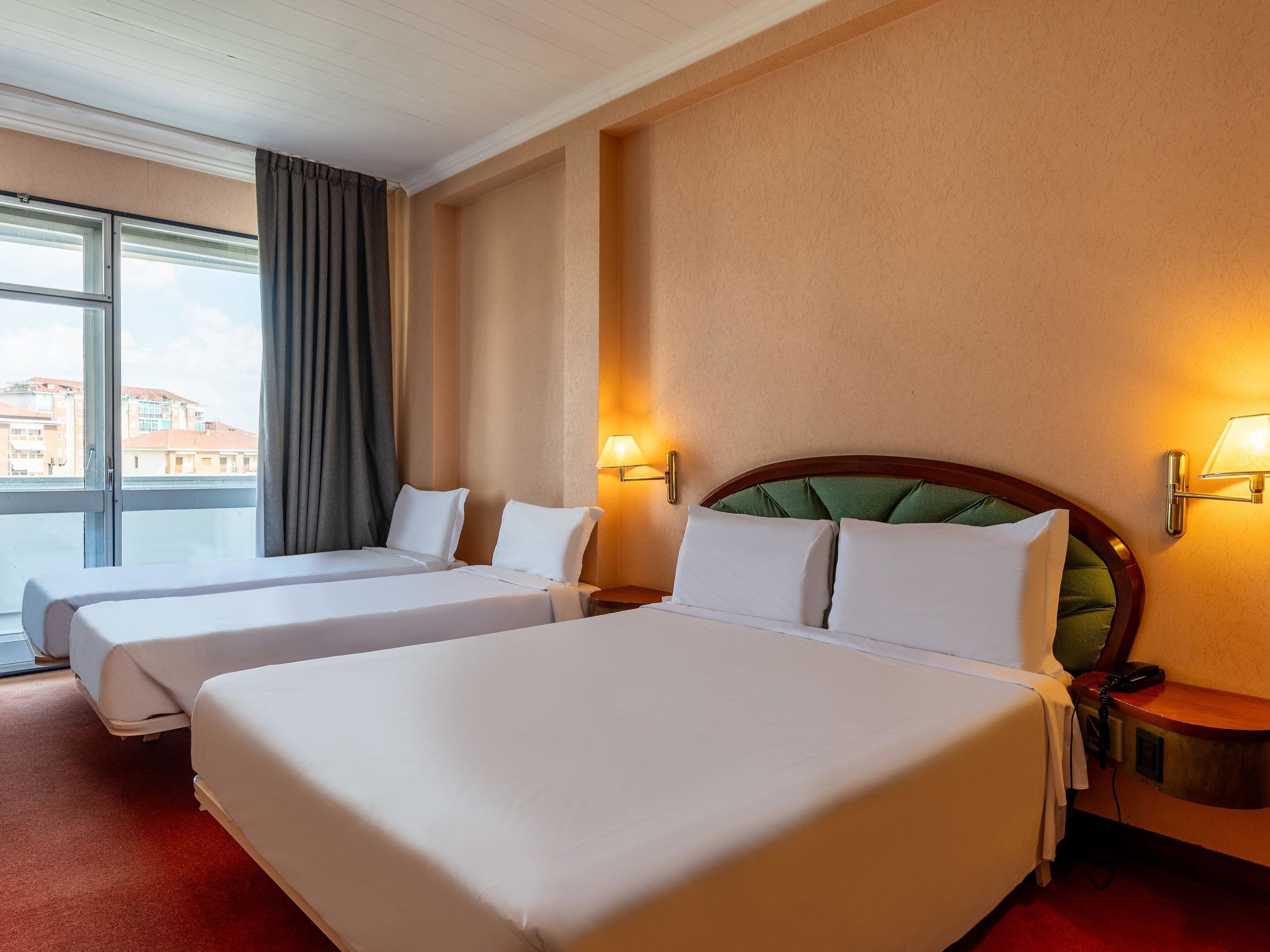 Habitación de hotel con dos camas, un ventanal con cortinas, una lámpara de pared y un teléfono en la mesita de noche.