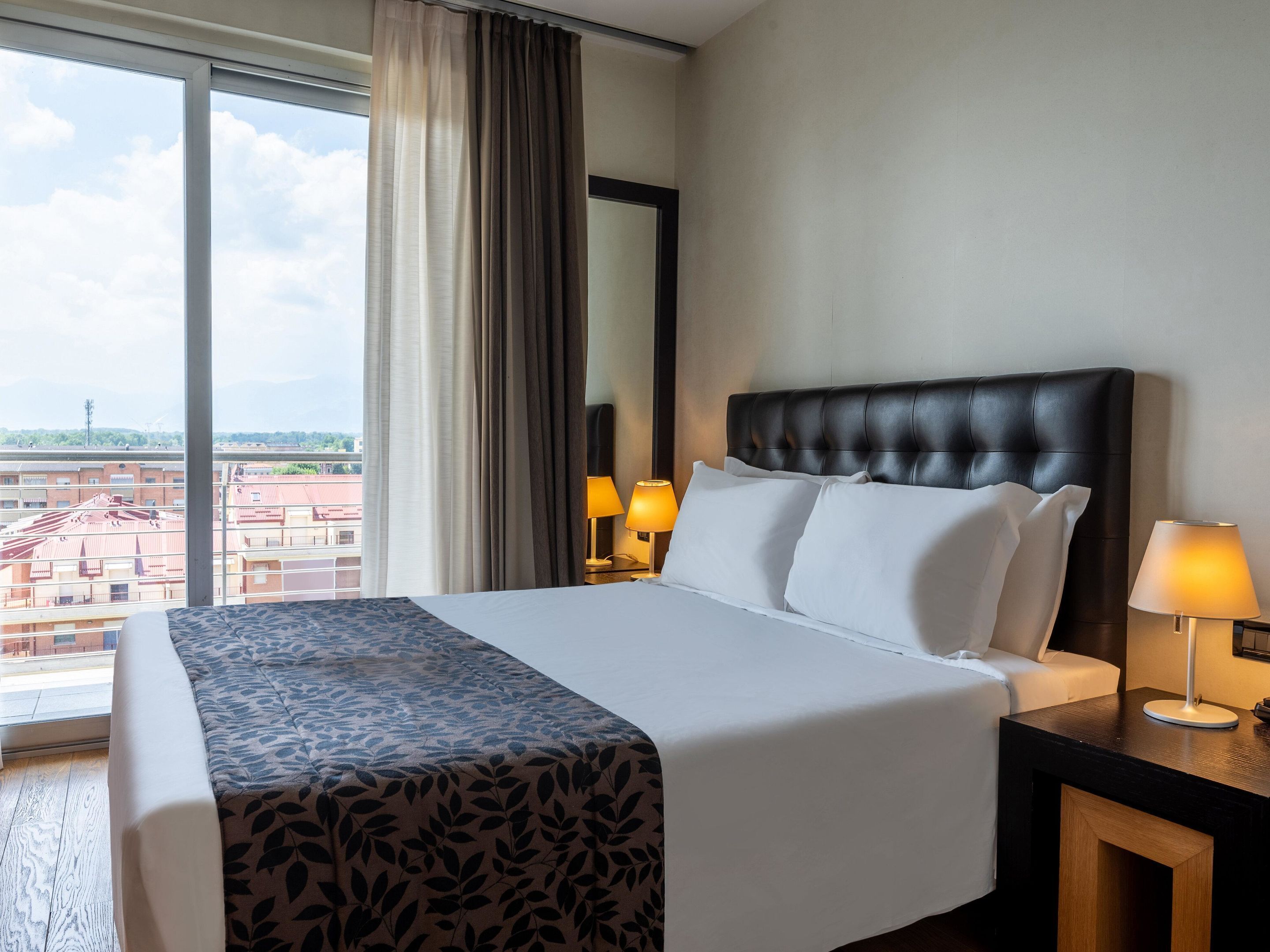 A hotel room with a large bed, two bedside lamps, a window with curtains, and a view of buildings.