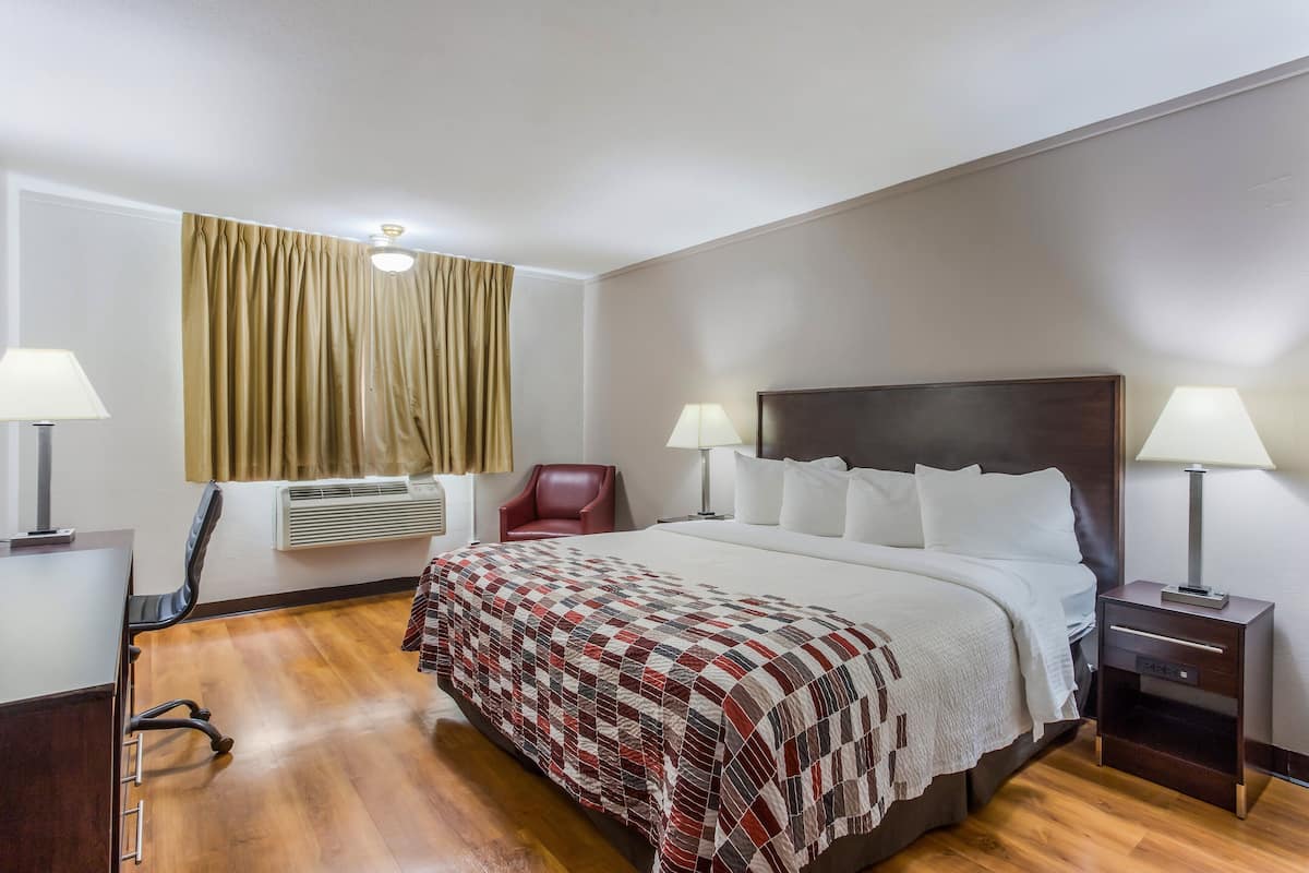 A hotel room with a large bed, two bedside tables with lamps, a red chair, a desk, and an air conditioning unit.