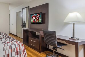 A hotel room with a bed, a desk with a chair, a television mounted on the wall, and a lamp on the desk.