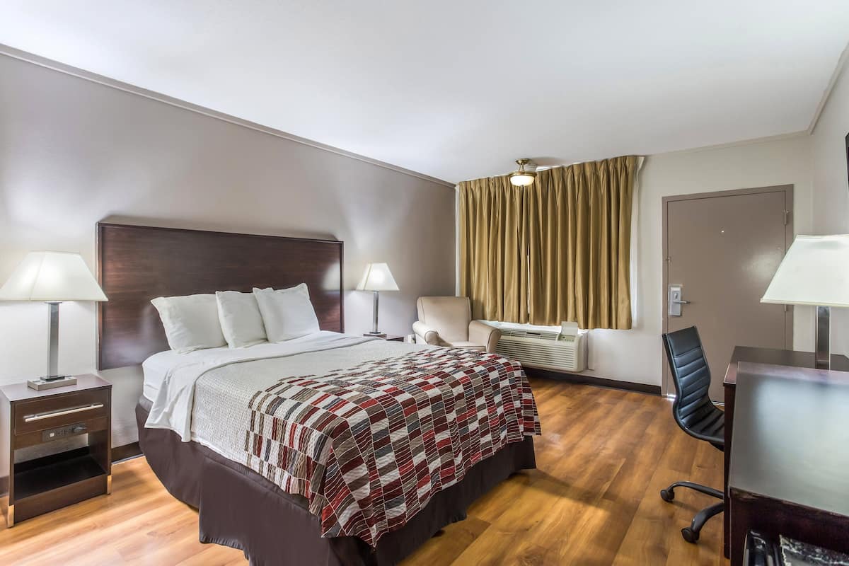 A hotel room with a large bed, two bedside tables with lamps, a desk with a chair, a window with curtains, and a door.