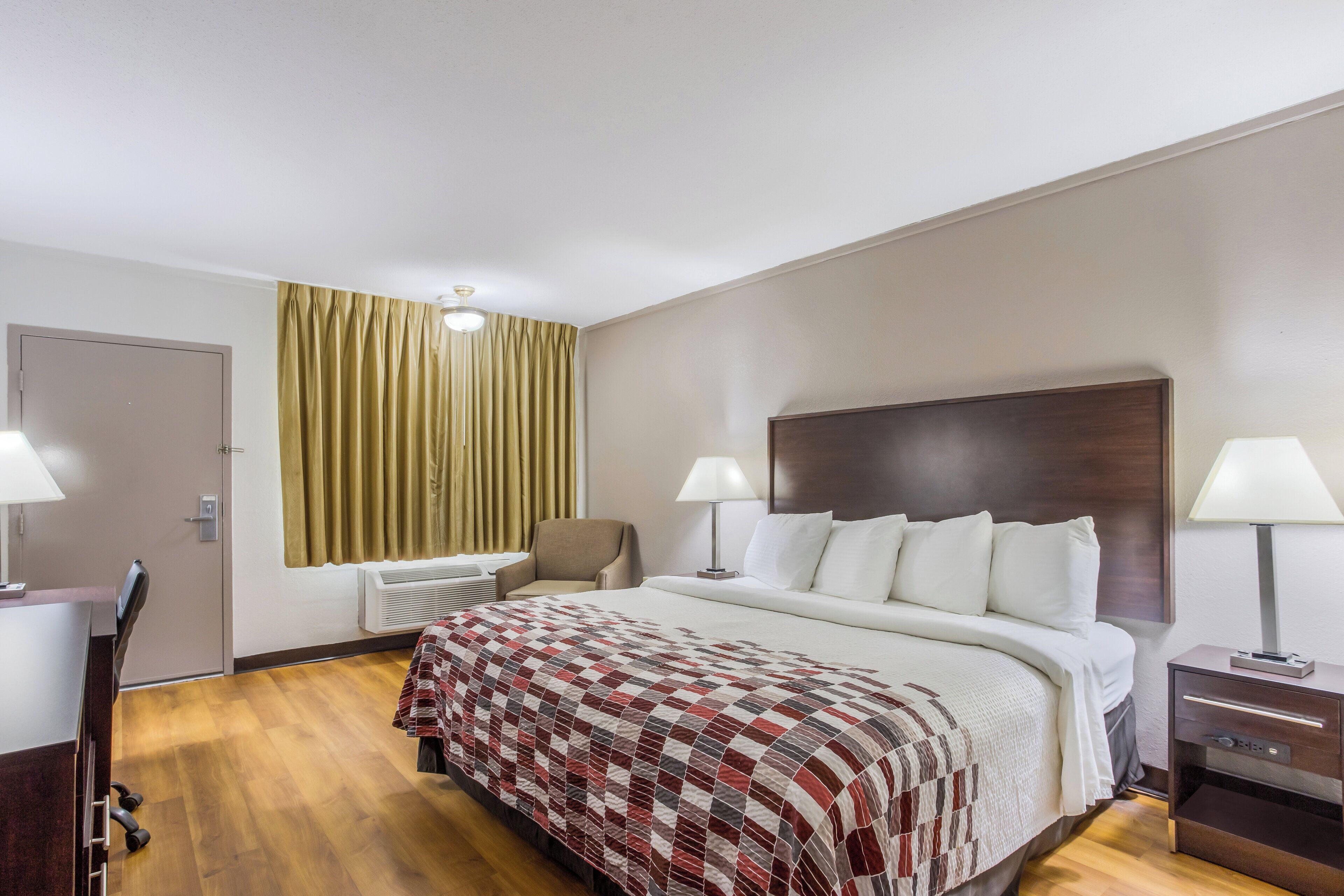 A hotel room with a bed, two bedside lamps, a chair, a desk, and a window with curtains.