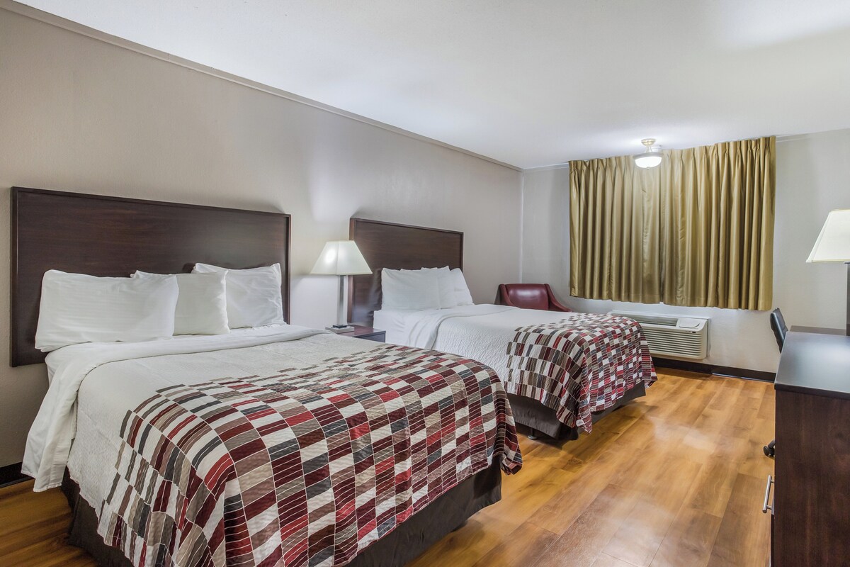 A hotel room with two beds, a wooden floor, a window with curtains, and a ceiling light.