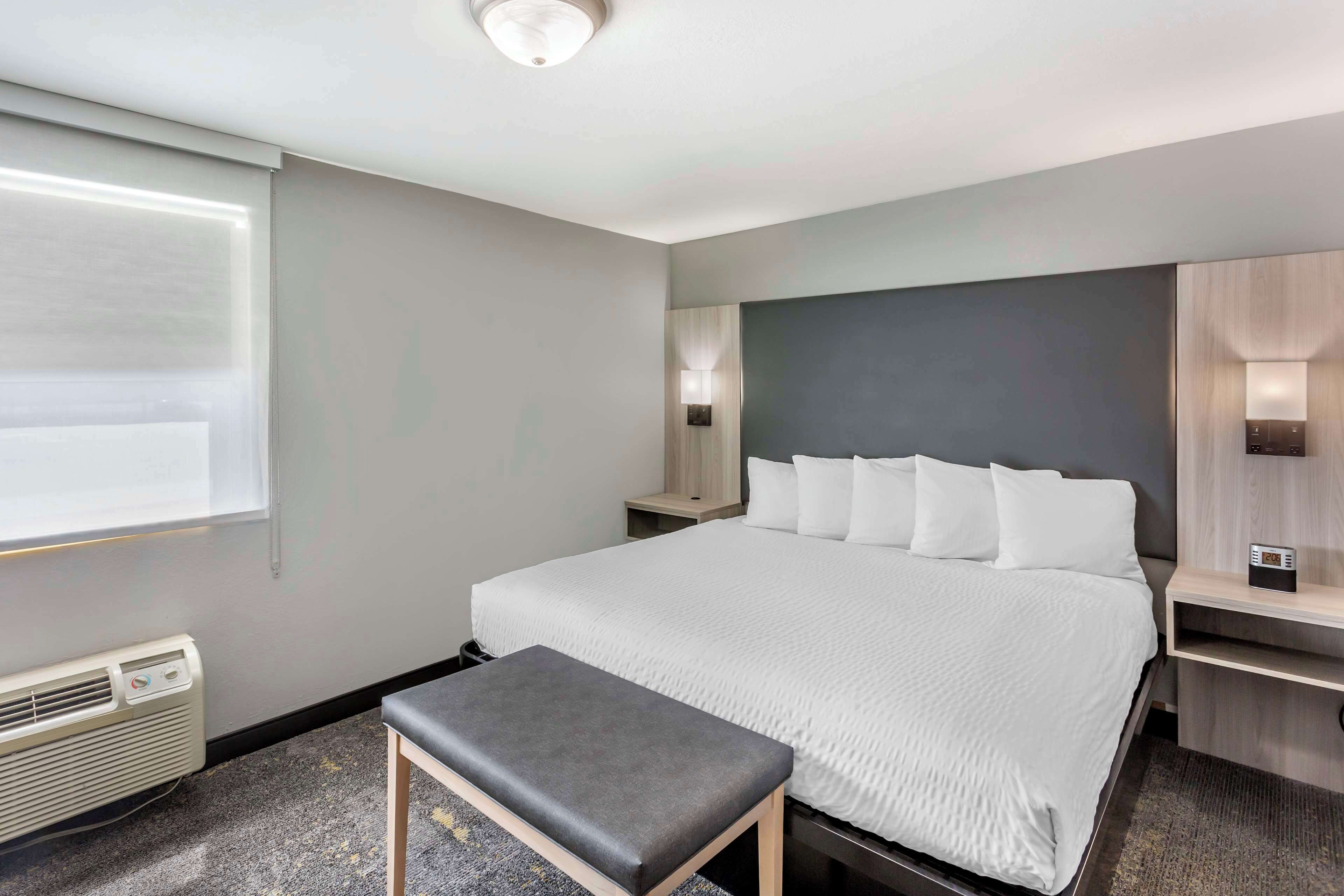 Una habitación de hotel moderna con una cama grande, una mesita de noche, un banco, un calentador y una ventana con persianas.