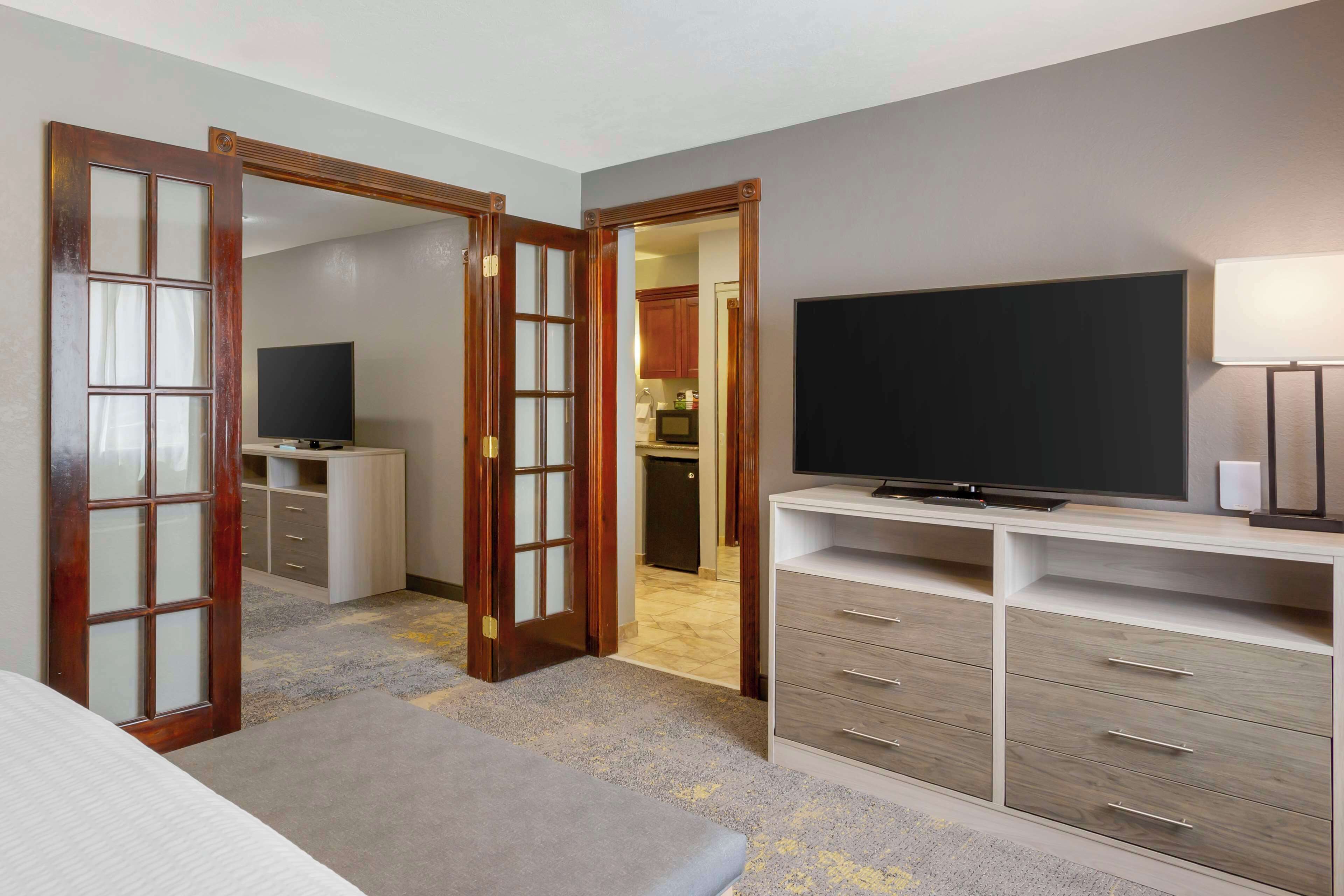 A hotel room with a bed, a TV, a desk with drawers, and a door leading to a kitchen.