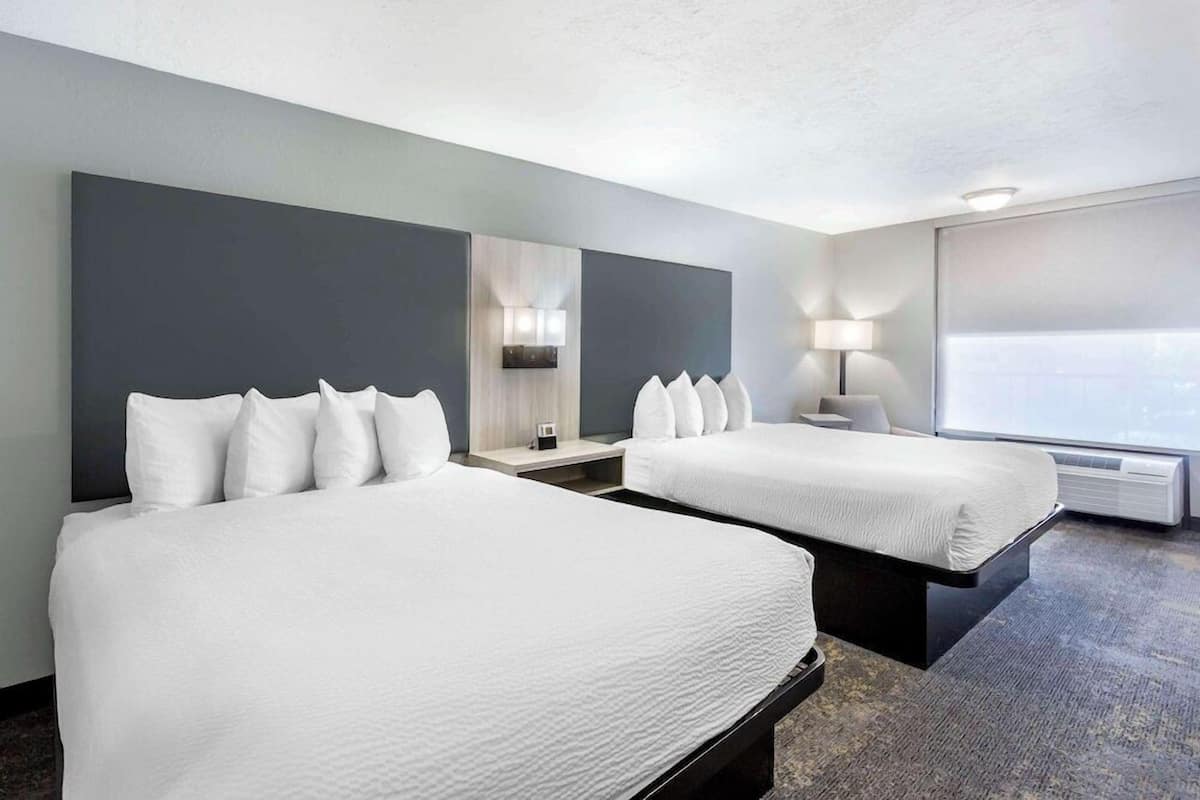 A hotel room with two beds, a large window, a wall-mounted TV, and a bedside table with a lamp.