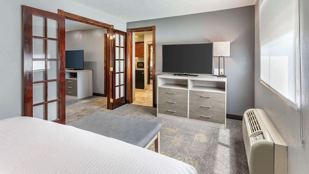 A hotel room with a bed, a desk, a television, a lamp, and a view of the kitchen. - Best Western Plus Weston Inn (Logan)