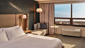 Une chambre d’hôtel comprenant un lit, un coin salon avec une table et une plante, une fenêtre donnant sur la ville et un radiateur.