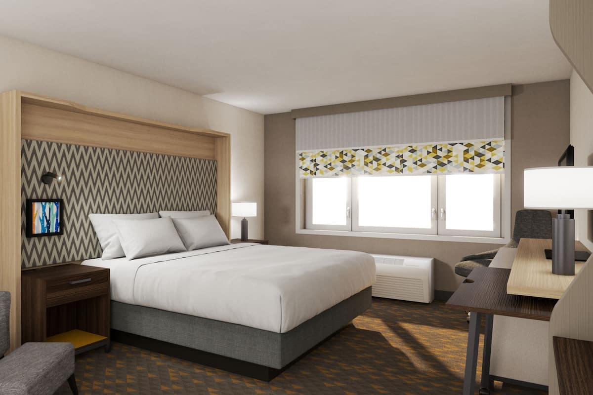 A hotel room with a bed, bedside tables, a desk, a chair, a window with curtains, and a wall-mounted TV.