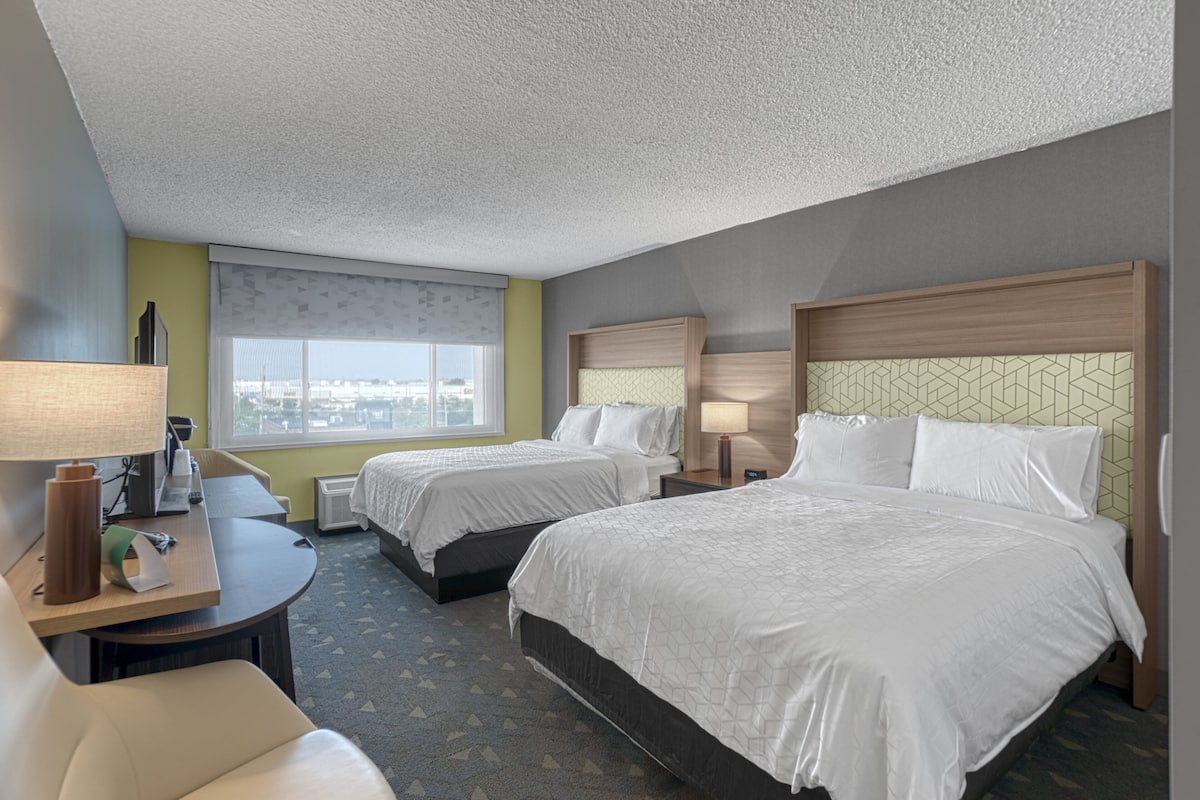 A hotel room with two beds, a desk, a chair, and a window with a city view.