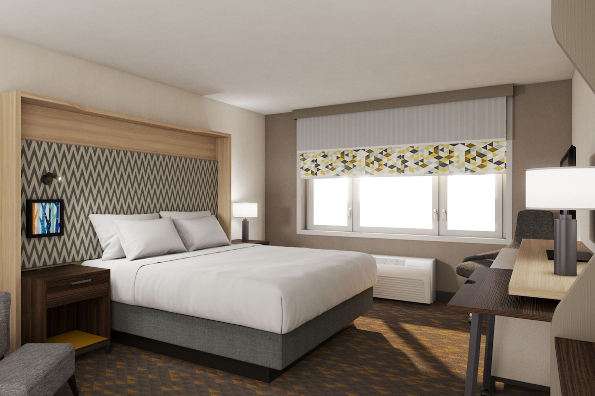 A hotel room with a bed, bedside tables, a desk, a chair, a window with curtains, and a wall-mounted TV.