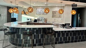 Un espace bar moderne avec un comptoir incurvé, de nombreux tabourets et des luminaires suspendus. Un tableau présentant le menu est visible sur le mur, et une machine à café se trouve sur le comptoir.