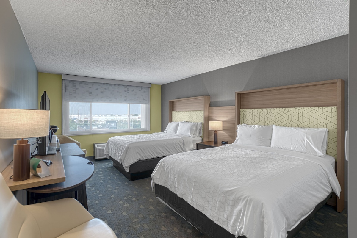 A hotel room with two beds, a desk, a chair, and a window with a city view.