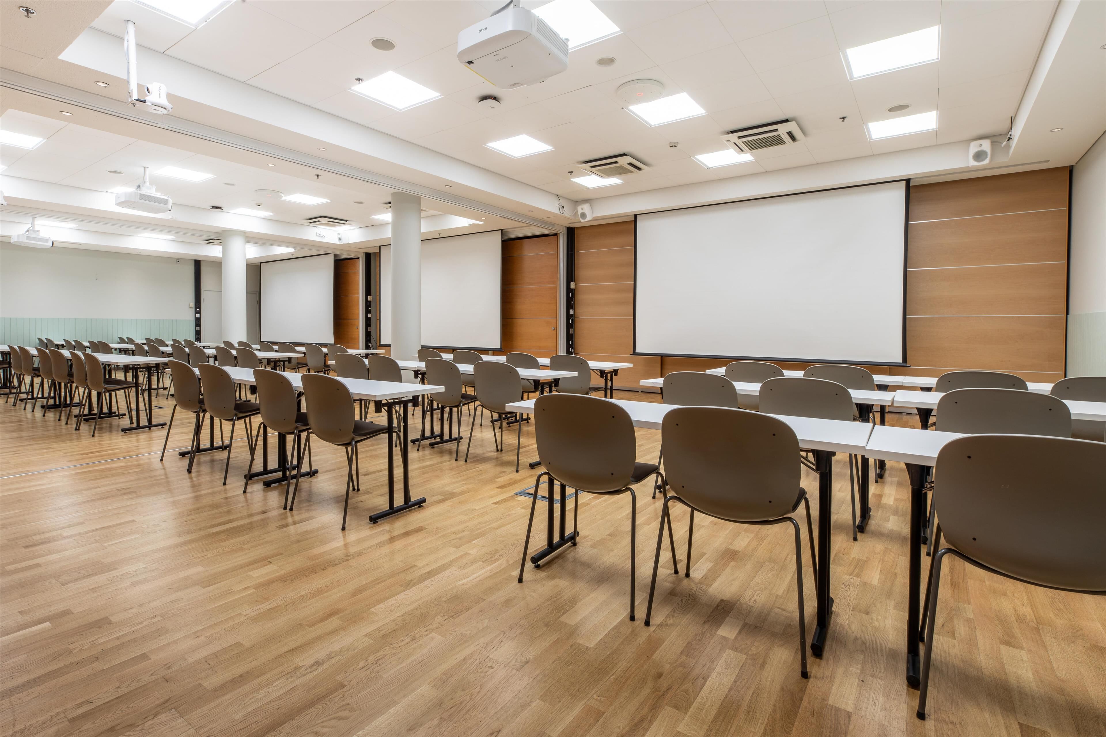 Un salón de conferencias con filas de mesas y sillas, un proyector y una pantalla grande.