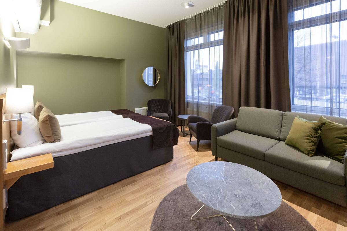 Ein Hotelzimmer mit Bett, Sofa, kleinem Tisch und Blick auf die Stadt durch große Fenster.
