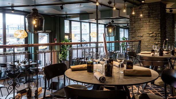 Ein Restaurant mit runden Tischen, schwarzen Stühlen, hängenden Leuchten und Blick auf die Stadt durch große Fenster.