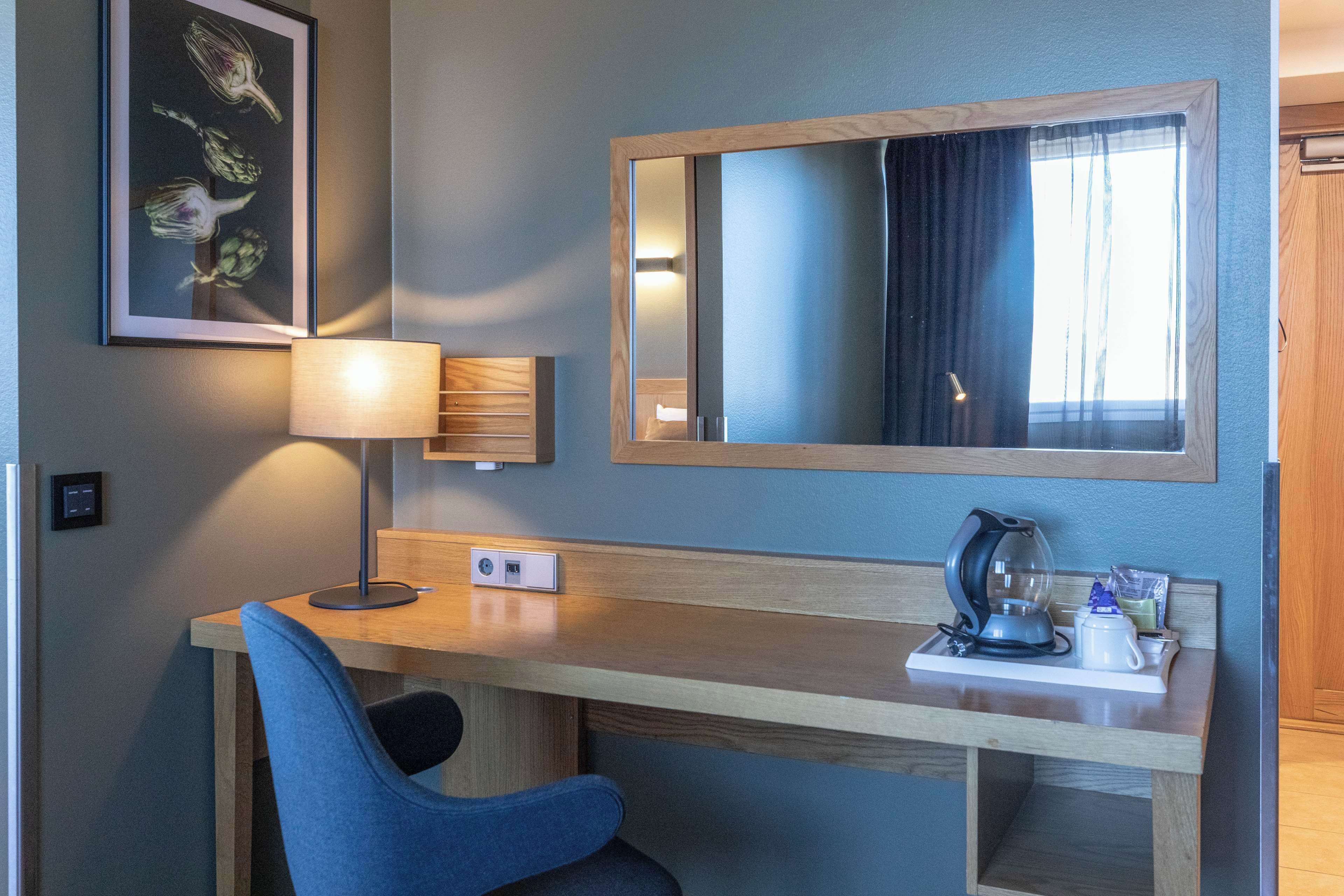Ein Hotelzimmer mit einem Holztisch, einem blauen Sessel, einer Lampe, einer Kaffeemaschine und einem gerahmten Bild an der Wand.