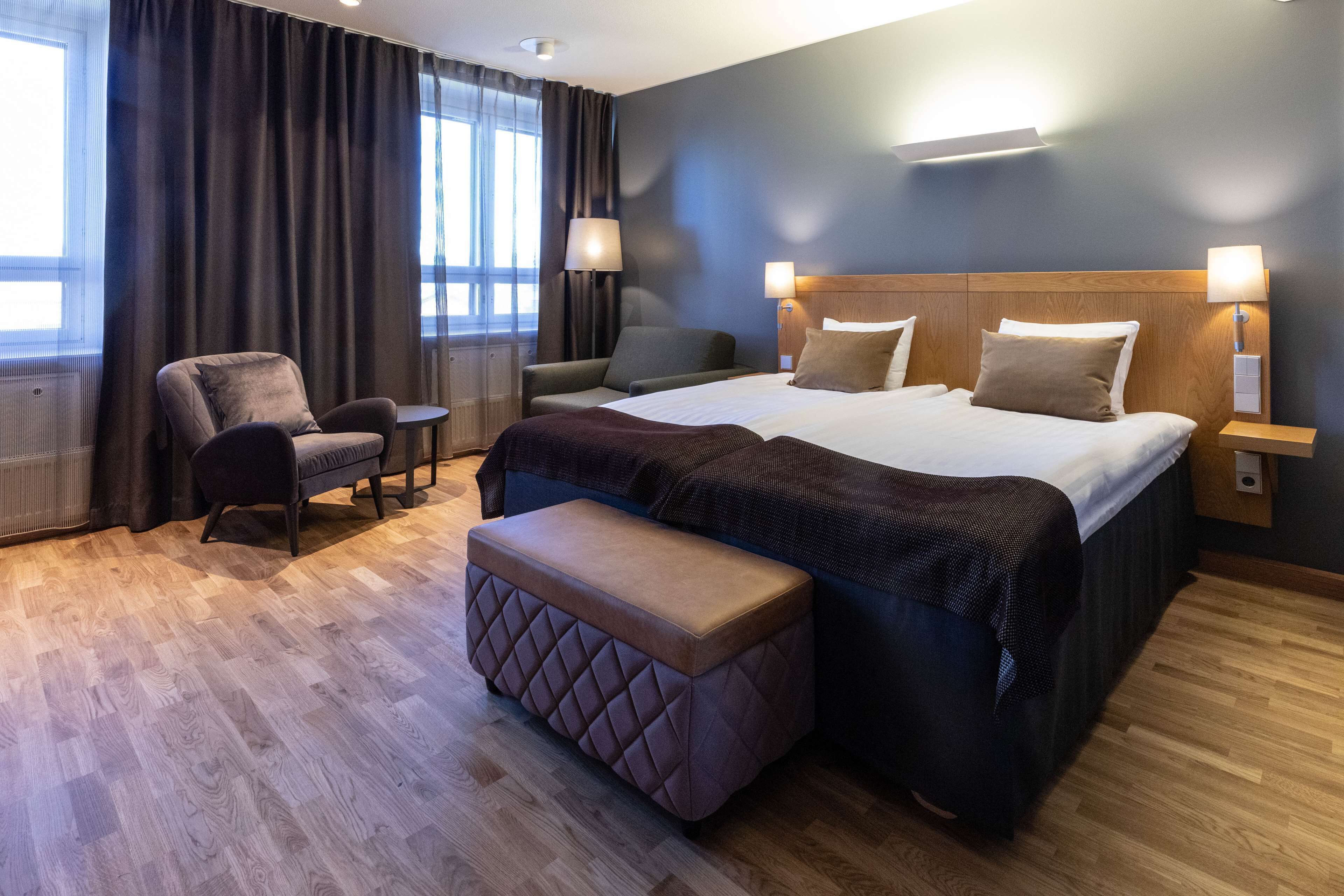 Una habitación de hotel con una cama grande, dos mesitas de noche, una silla, una pequeña mesa y un banco.