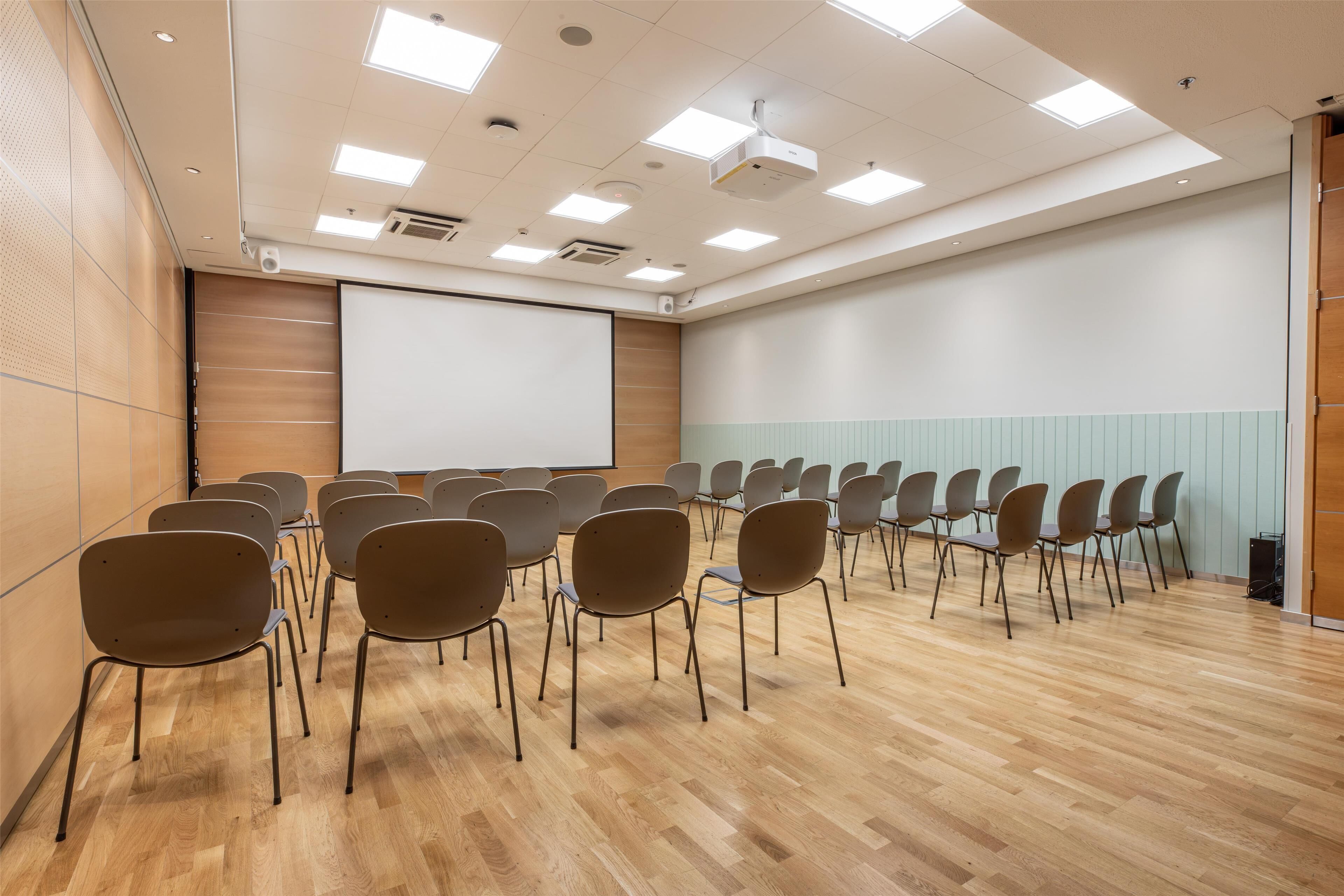 Un salón de conferencias con filas de sillas, una pantalla para proyector y suelo de madera.