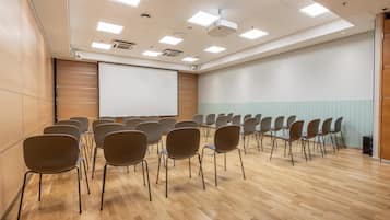 Un salón de conferencias con filas de sillas, una pantalla para proyector y suelo de madera.
