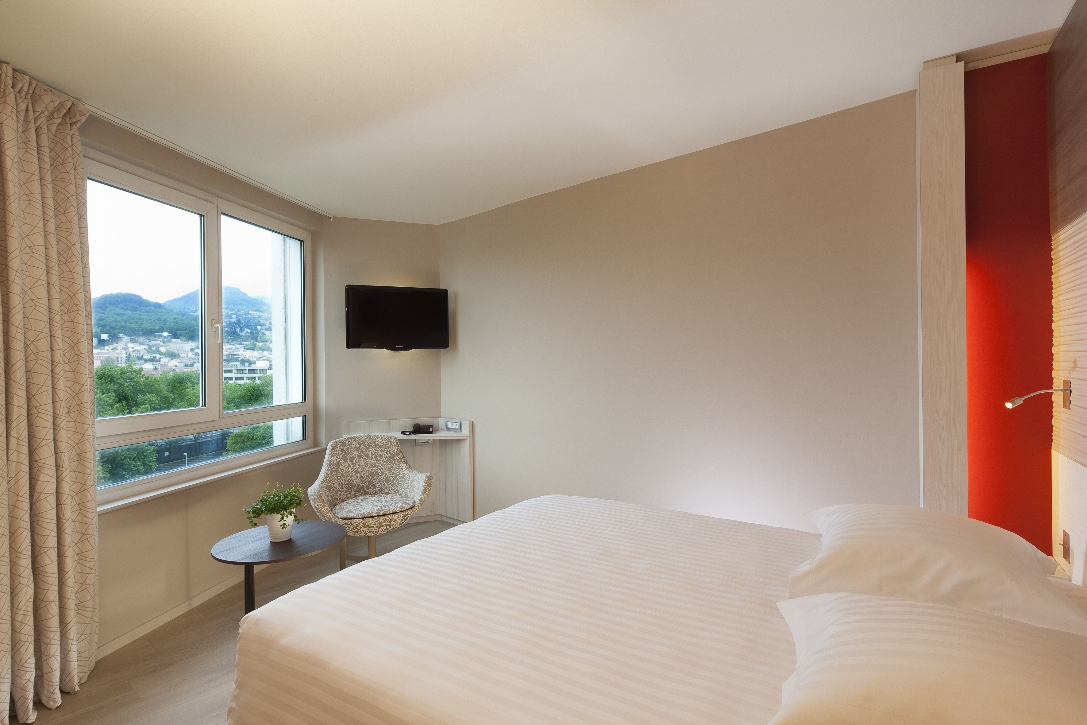 A hotel room with a large bed, a chair, a small table with a plant, a TV mounted on the wall, and a window with a view of mountains and a city.