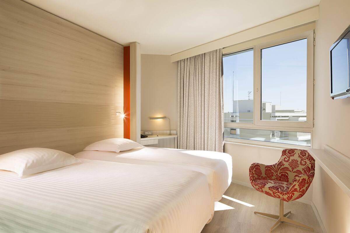 A hotel room with two beds, a window with curtains, a desk with a lamp, and a red patterned chair.