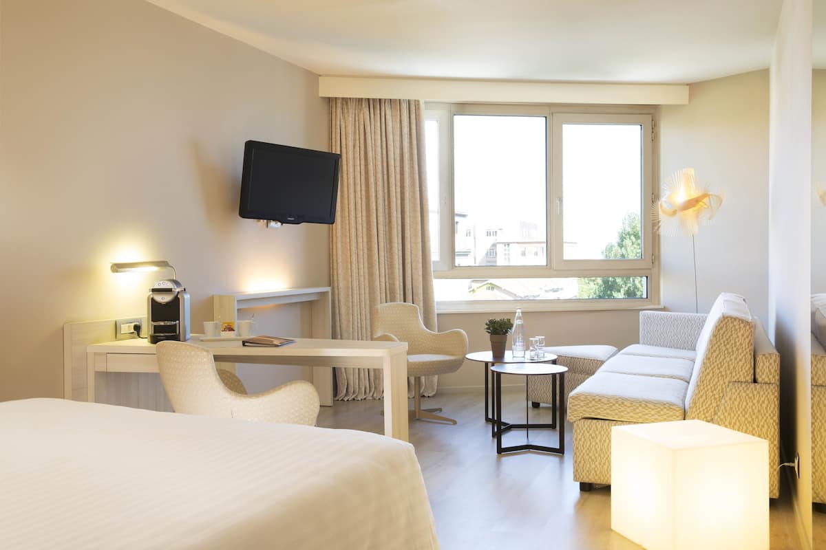 A hotel room with a bed, a desk with a coffee maker, a TV, a sofa, a small table with a plant, and a window with curtains.