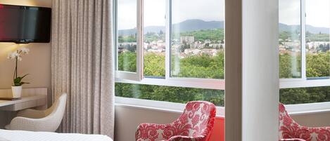 A hotel room with a bed, two red patterned chairs, a TV, and a large window offering a city view.