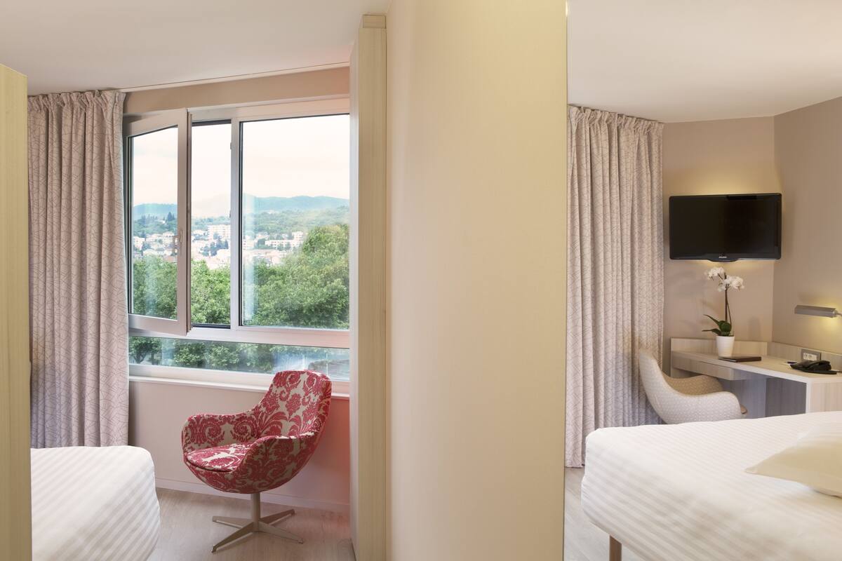 A hotel room with a bed, a desk with a flat-screen TV, a chair, and a large window offering a city view.