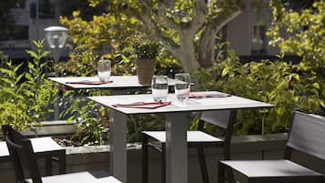 Un espacio exterior para comer con mesas y sillas, rodeado de follaje y un edificio al fondo.