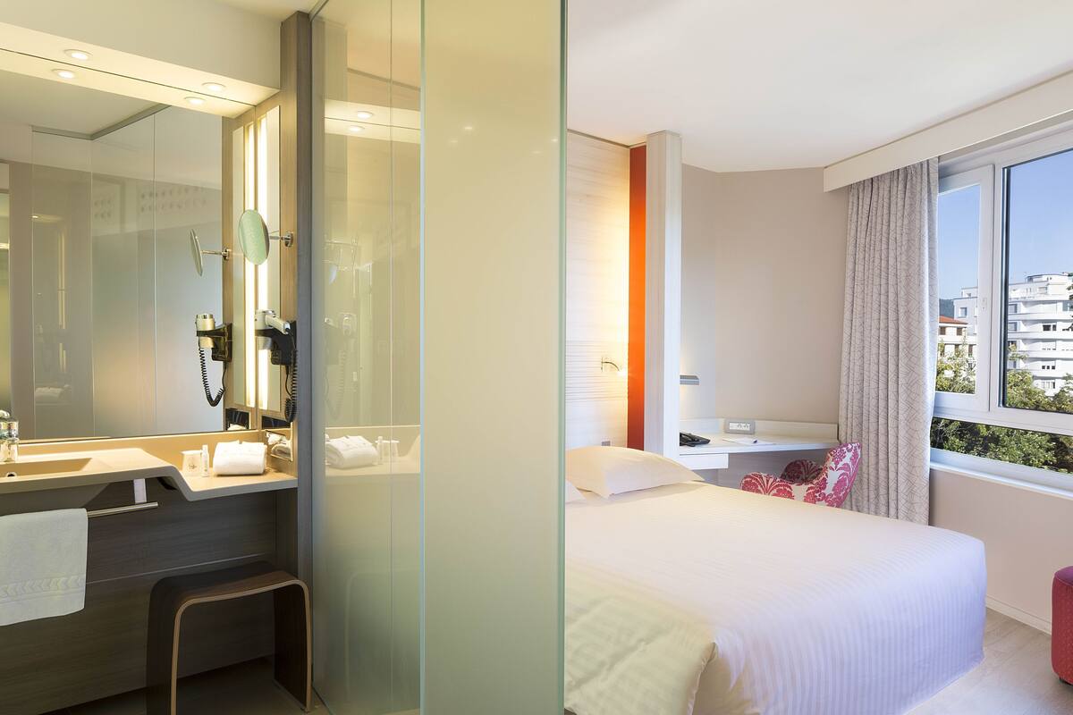A modern hotel room with a glass partition, a bathroom with a sink and mirror, a bed with pillows, and a window with a view.