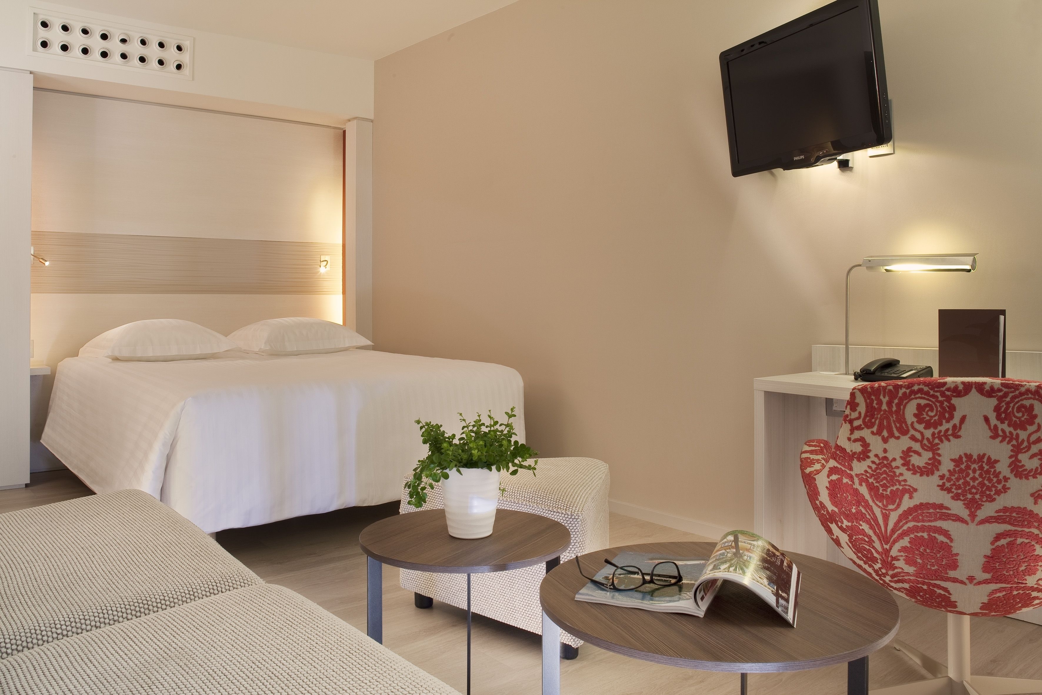 A hotel room with a bed, a small round table with a plant, a red patterned chair, a sofa, and a TV mounted on the wall.