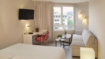 A hotel room with a bed, a desk with a chair, a sofa, a TV mounted on the wall, and a window with a view of buildings.