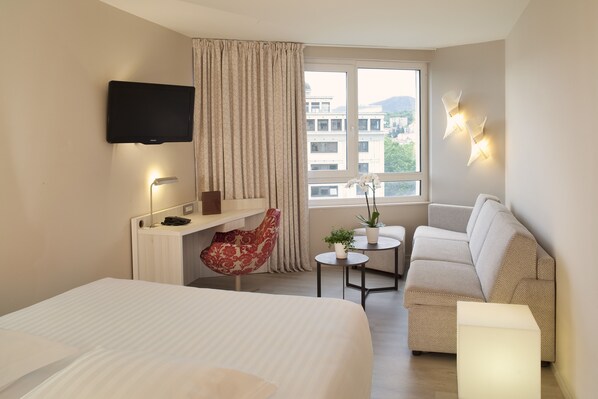Habitación de hotel con cama, escritorio con silla, sofá, televisión montada en la pared y ventana con vista a edificios.