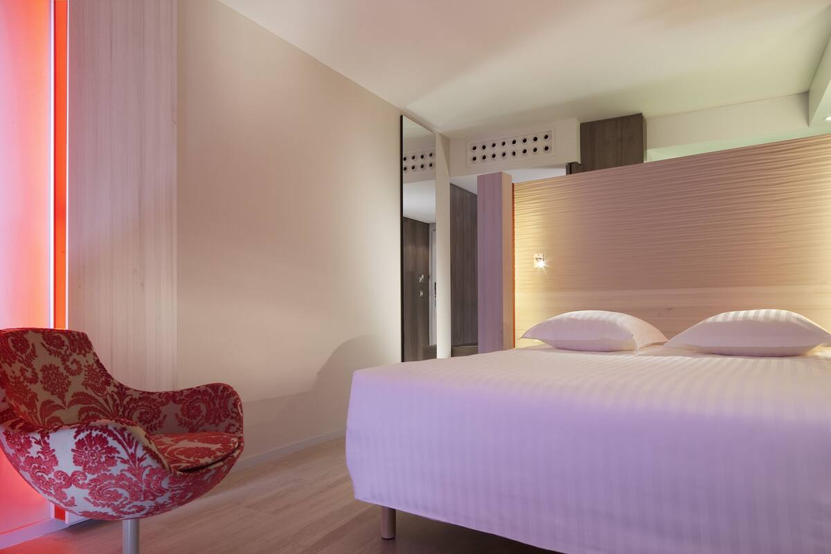 A modern hotel room with a bed, a red upholstered chair, and a wooden headboard.