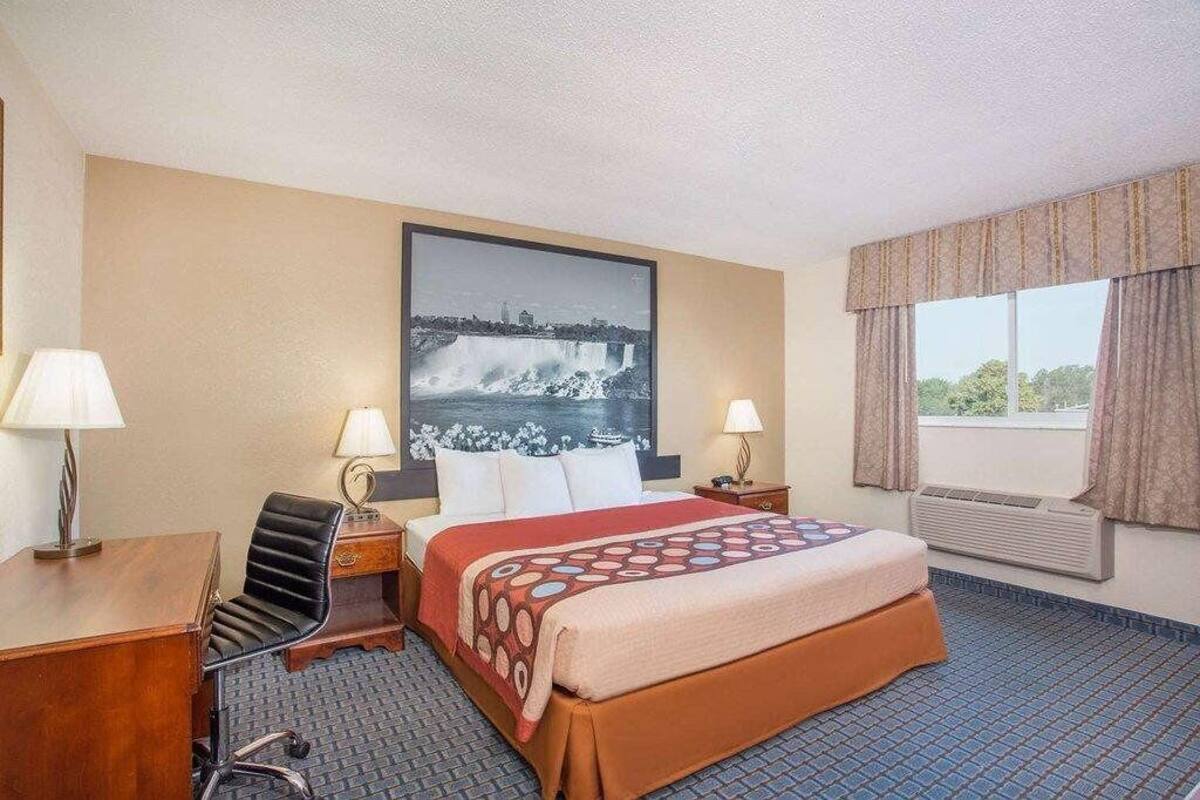 A hotel room with a bed, two bedside tables, a desk, a chair, a window with curtains, and a wall-mounted picture.