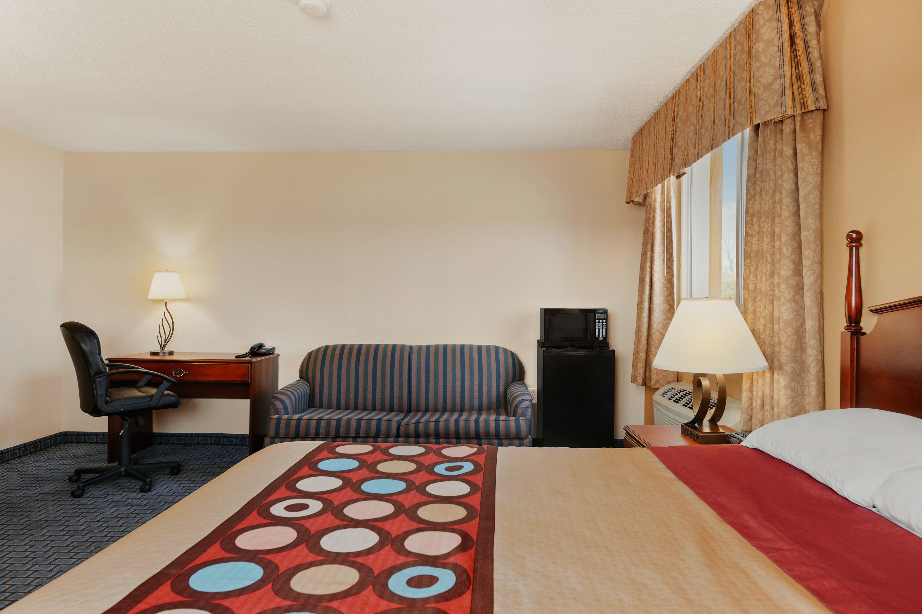 Habitación de hotel con cama, escritorio con lámpara, silla, sofá, televisión y ventana con cortinas.